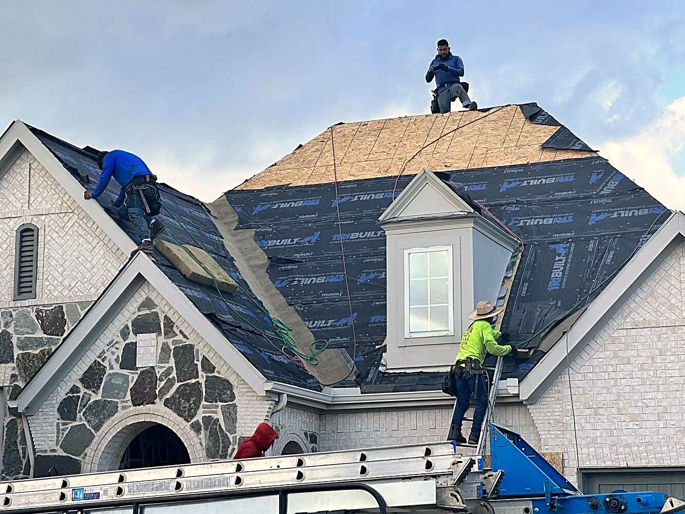 Roofers laying synthetic underlayment over the deck of a stone-front home